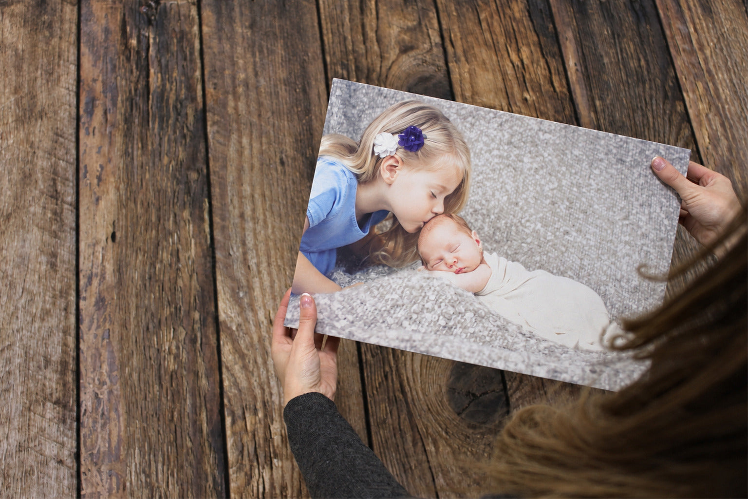 Hands holding a canvas print of a child kissing a newborn above a dark wooden table.