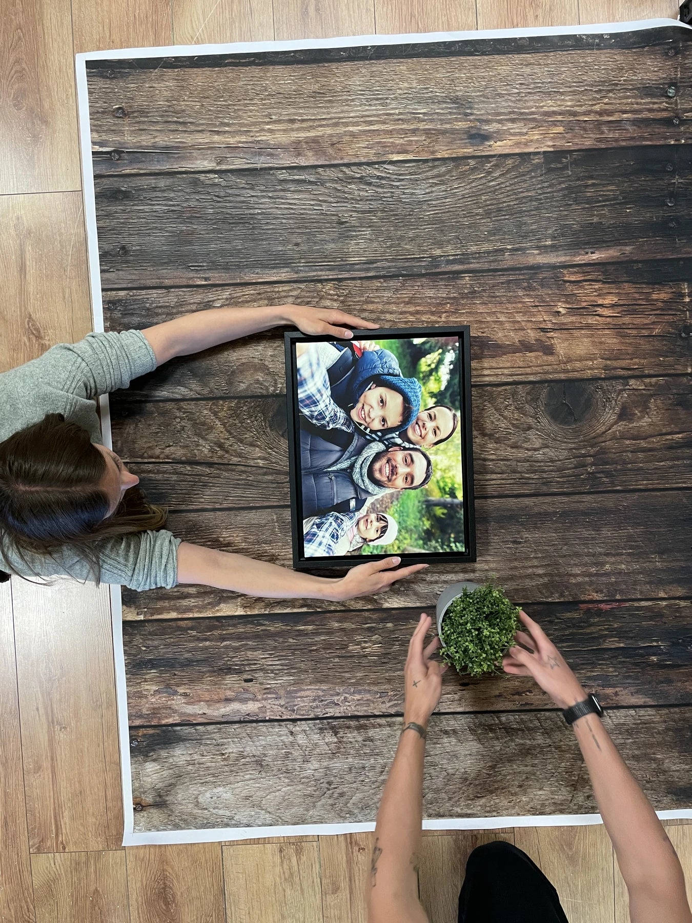 Complete workshop setup with two people arranging a framed family print, plant and wood-effect paper backdrop