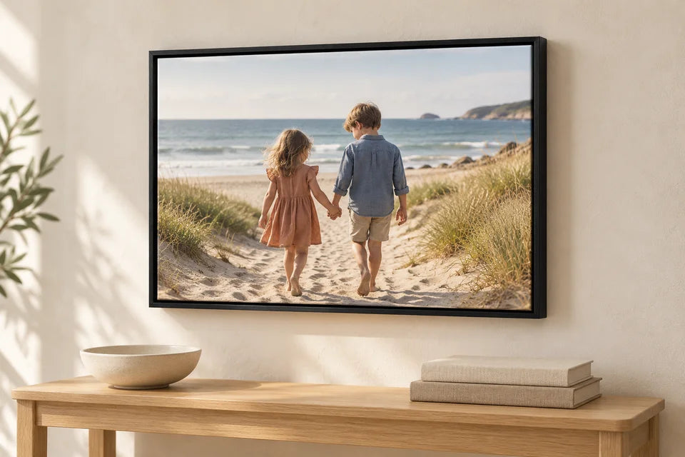 Two children on a beach printed on a canvas in a black floating frame, above a small oak console.