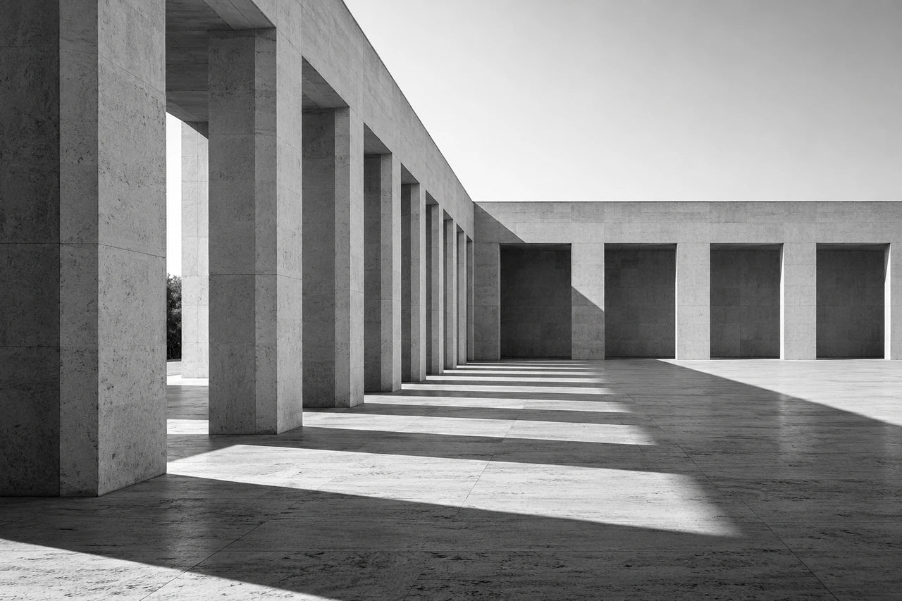 A fictional monochrome concrete colonnade and courtyard with repeated pillars and long geometric shadows.