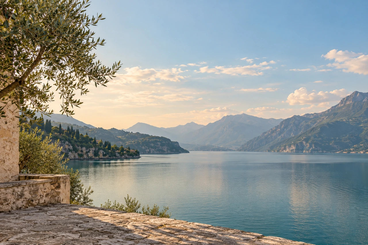 A fictional photographic-style lake view framed by olive branches and pale Mediterranean stone.