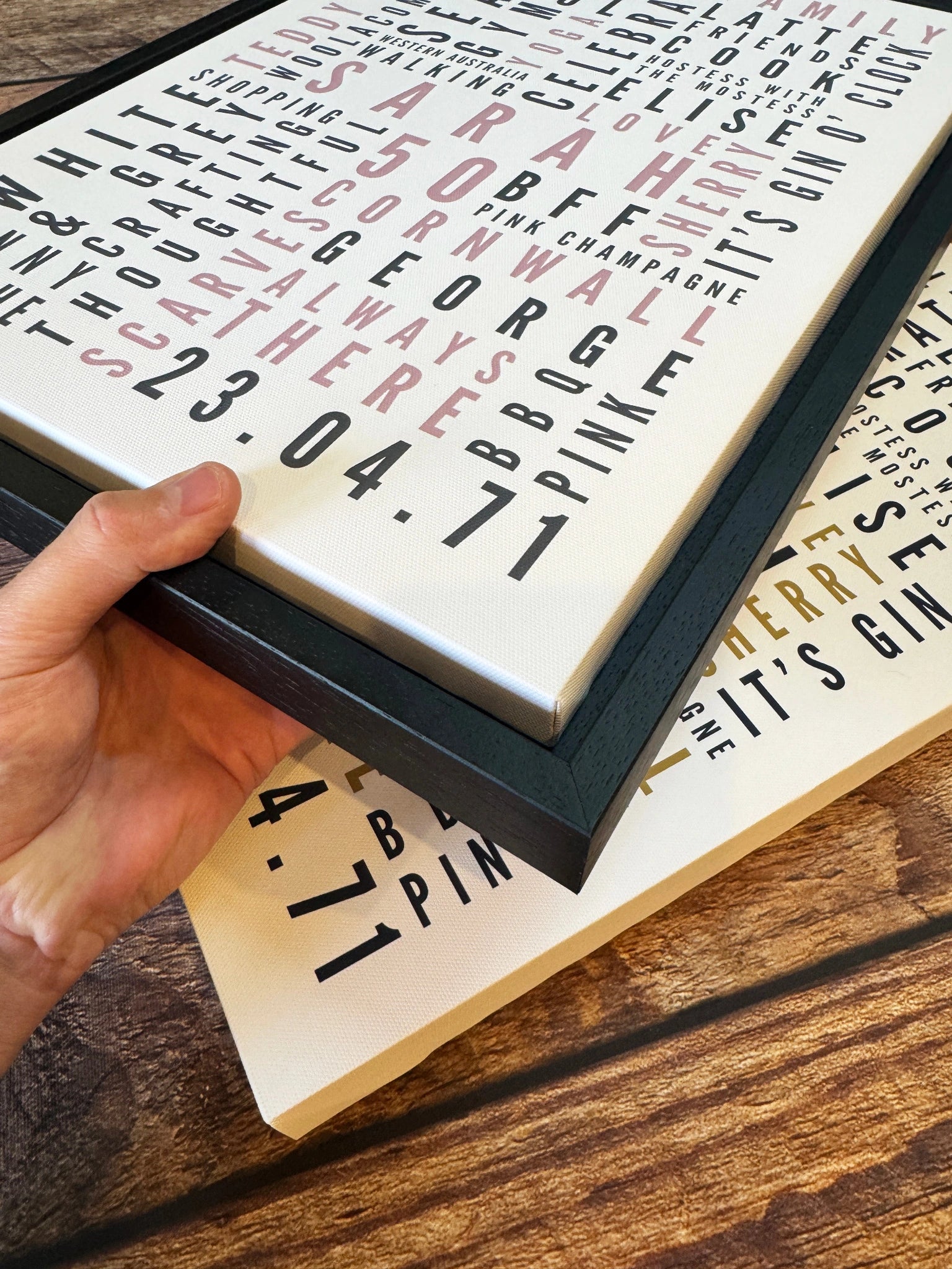 A hand holding a black floating frame around a personalised Word Art canvas.