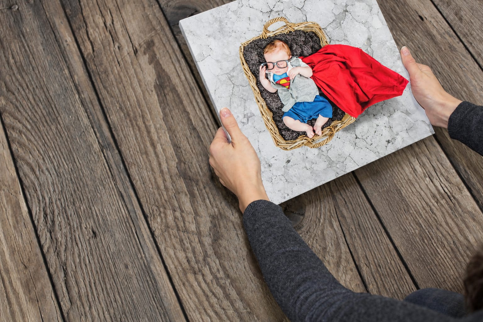A genuine photo canvas of a baby in a superhero outfit, held over a wooden table