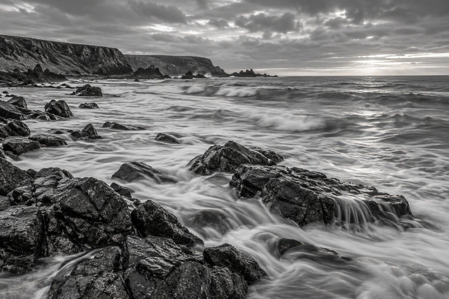 Monochrome rocky coastline with foaming water, low cliffs and a luminous cloudy horizon.