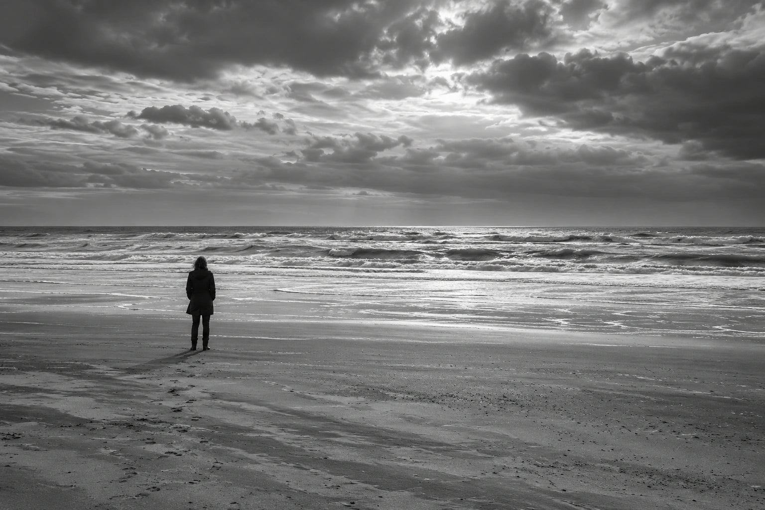 One distant person on a broad windswept beach facing an expansive sea beneath a dramatic clouded sky.