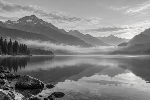 Black-and-white mountain lake with mist between distant hills and a quiet shoreline.