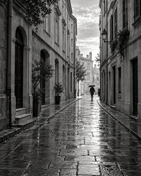 Black-and-white stone city street after rain, with wet paving and a distant umbrella.