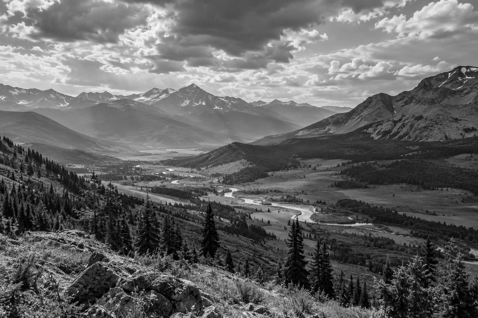 Black-and-white mountain valley with a winding river, layered slopes and moving cloud shadows.