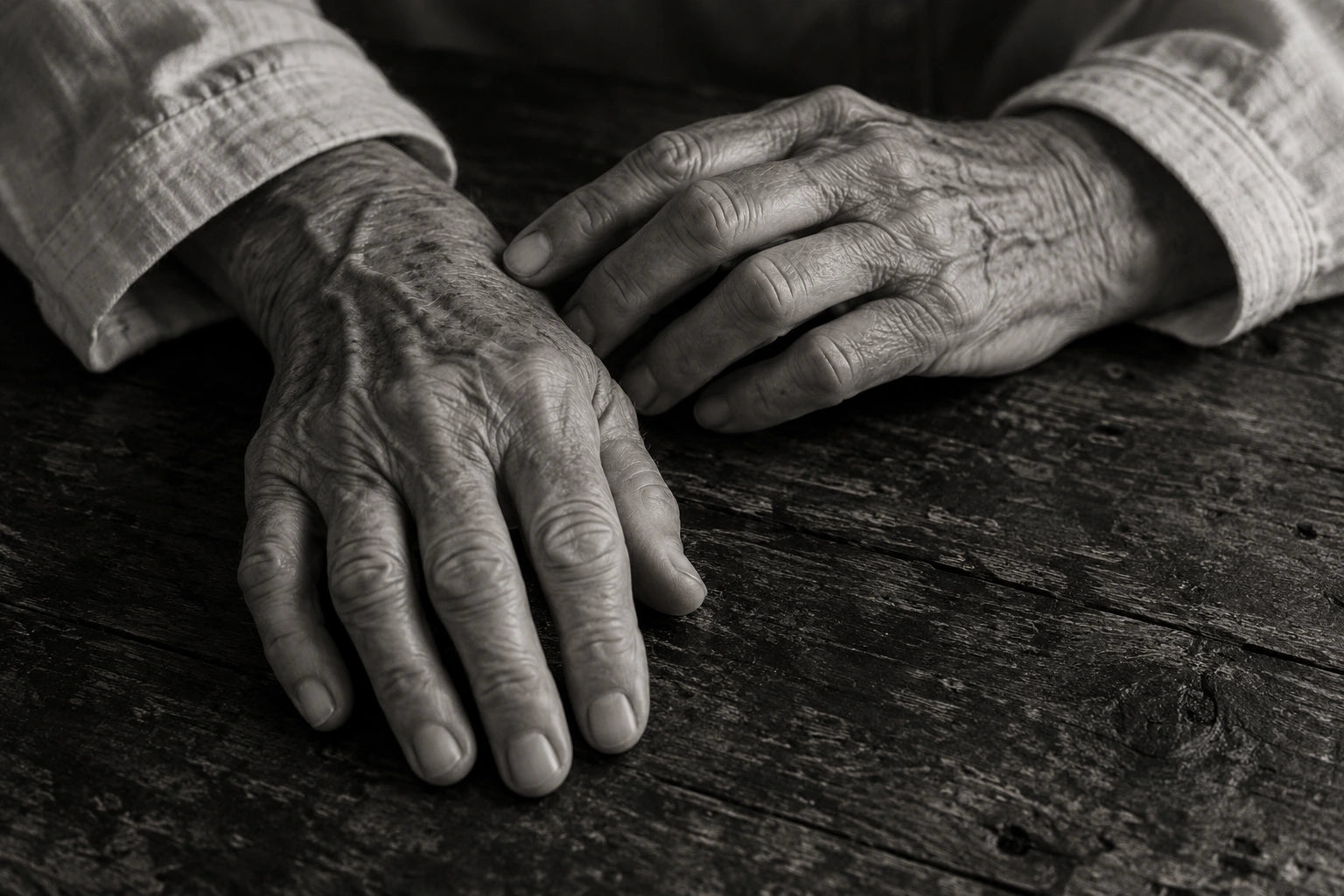 Two older hands resting naturally on a worn wooden table, lit from the side, with a plain linen cuff.