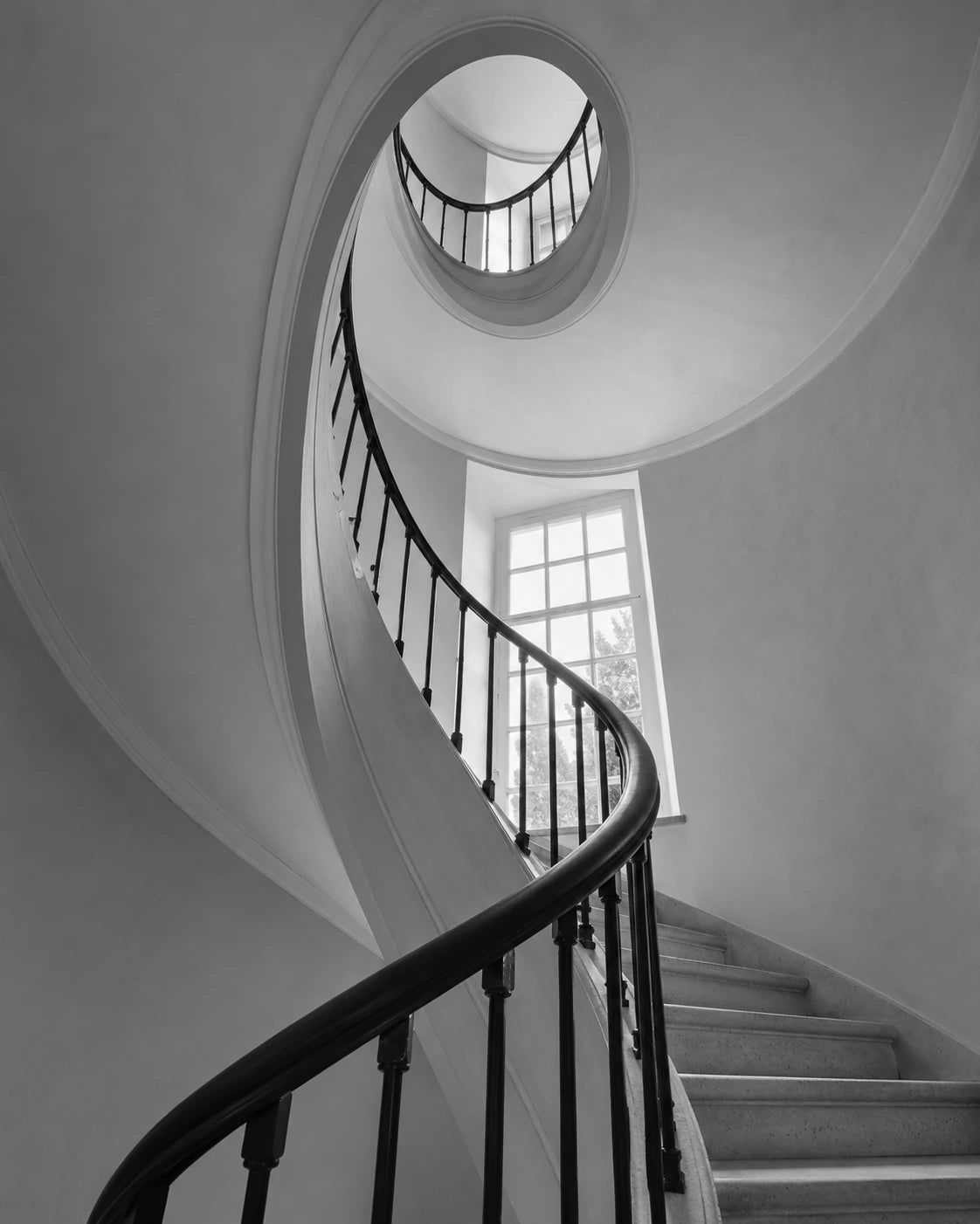 Black-and-white view up a curved staircase with white walls, dark rail and soft window light.