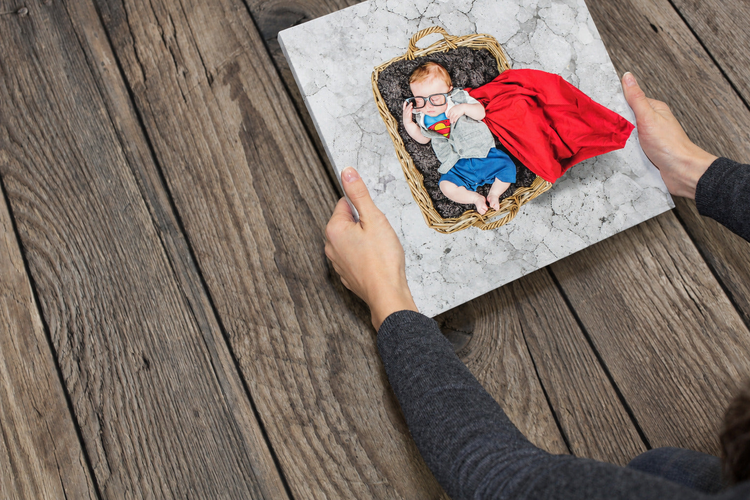 Hands holding a superhero baby canvas print above a dark wooden table.