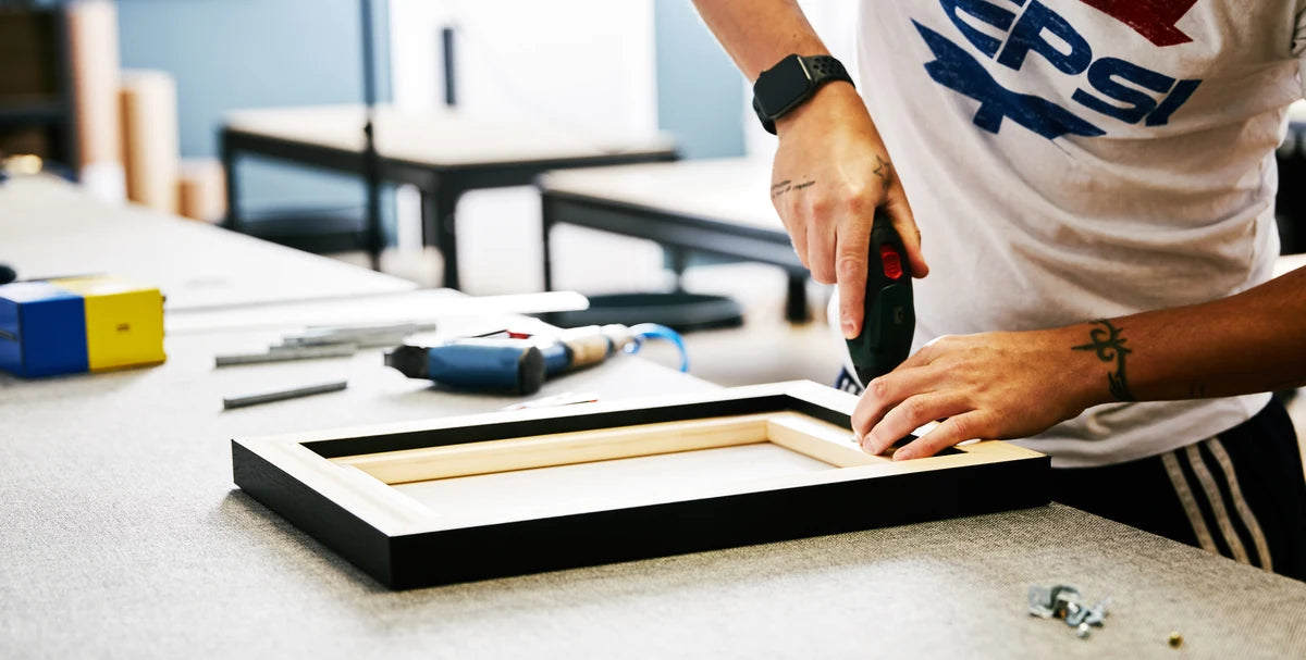 Hands working on the back of a black floating frame at the YI2C workshop bench.
