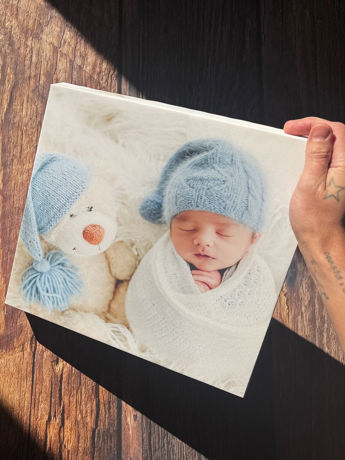 A newborn and teddy bear photograph printed on canvas, held above a dark wooden table.