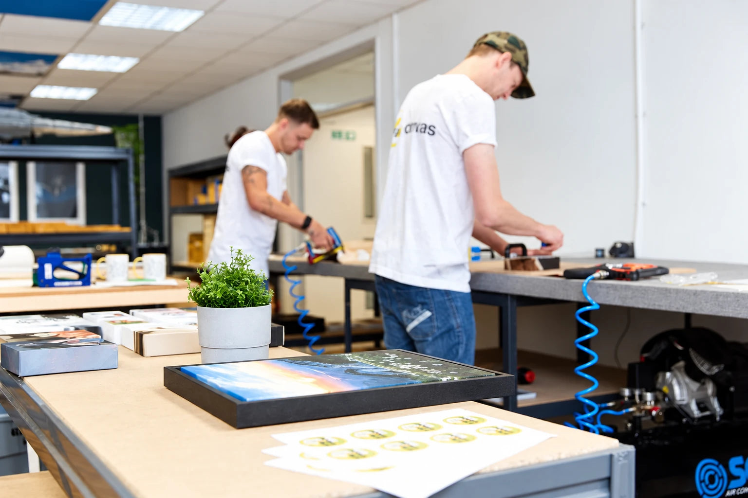 Two team members working at canvas-printing workshop benches, with a framed print on the foreground table.